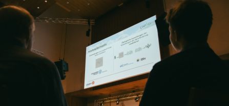 Große Leinwand mit Präsentation, im Vordergrund Zuhörer
