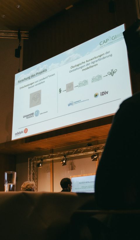 Große Leinwand mit Präsentation, im Vordergrund Zuhörer