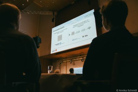 Große Leinwand mit Präsentation, im Vordergrund Zuhörer